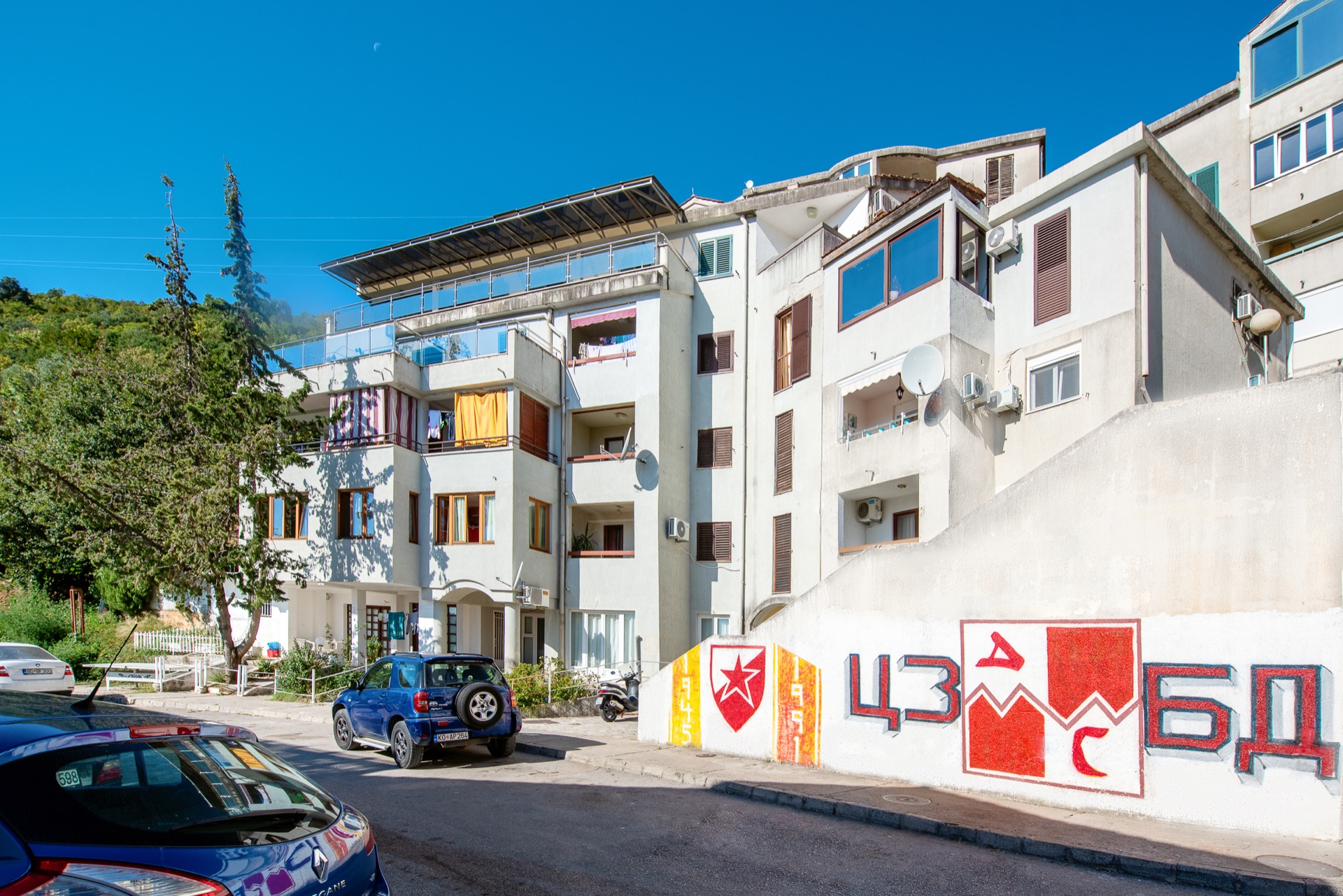 Facade of an apartment building in Budva, Montenegro, with balconies, terraces, and graffiti on the retaining wall by the road.