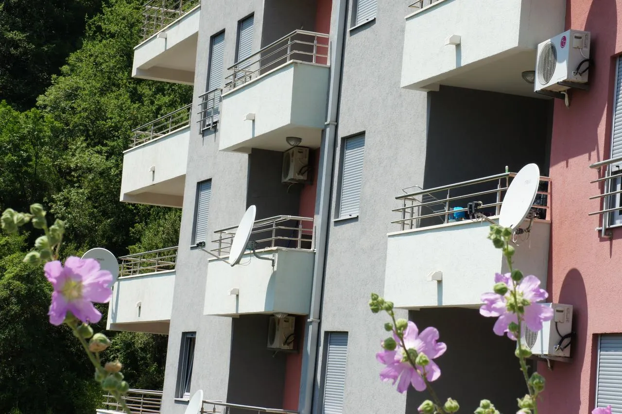 Exterior facade of a multi-story apartment building featuring balconies, AC units, and satellite dishes against greenery.