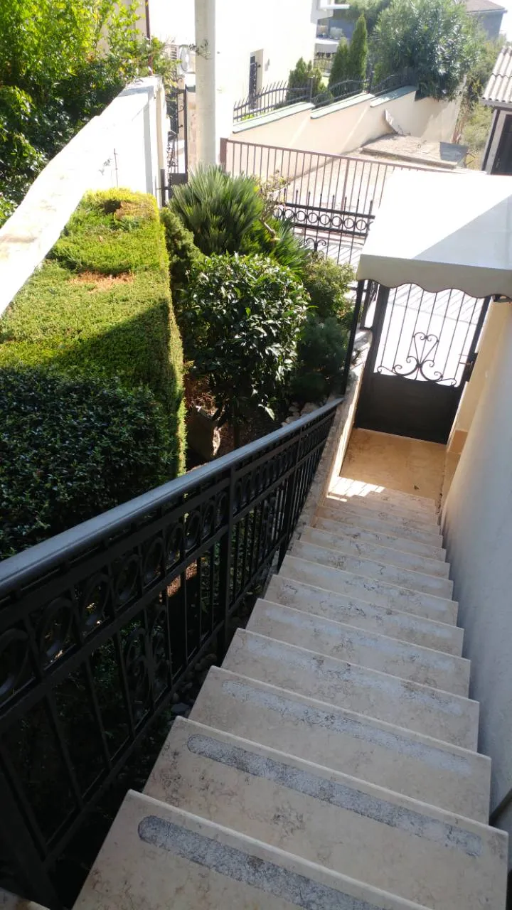 Stone staircase with black wrought iron railing leading down to a garden area with hedges and trees next to a gate.