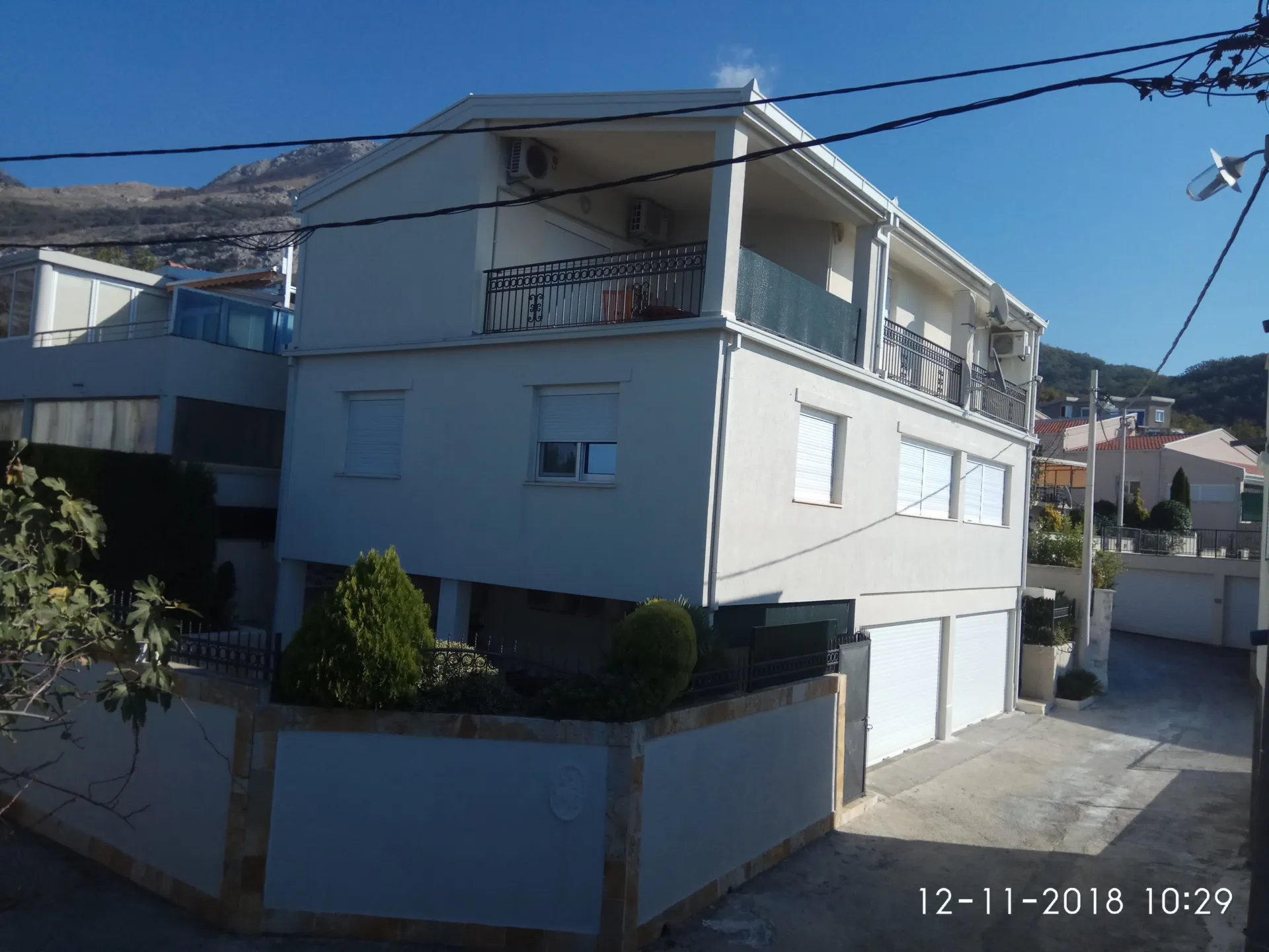 Exterior view of a two-story white house with balconies, garage doors, and a mountain background under a clear blue sky.