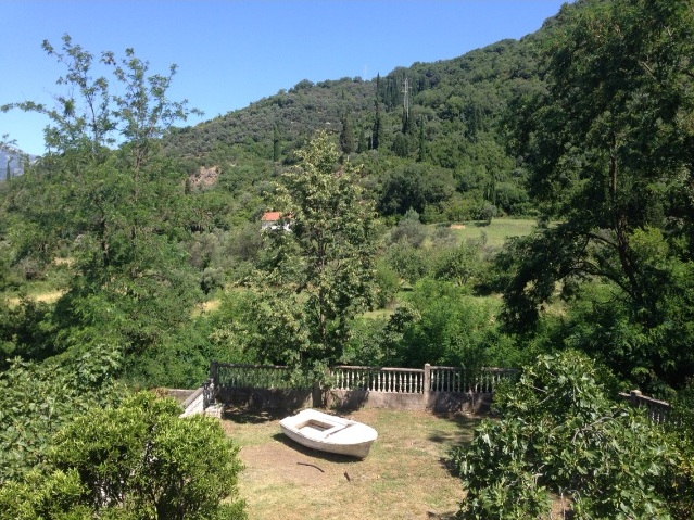 View of a green garden with a boat and mountains by the house in Tivat, Montenegro. Plot with a balustrade and dense vegetation.