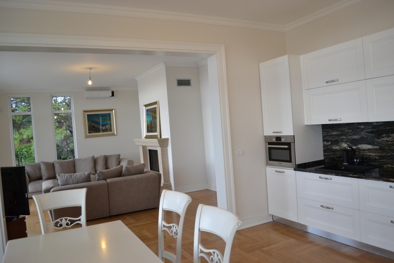 Open kitchen with white cabinets and dining area in foreground; living room with sofa and fireplace visible through archway.