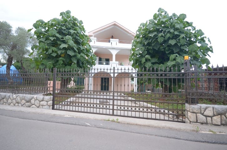 Three-story house with terraces and a wrought-iron fence in Krashichi, Montenegro, surrounded by green trees.