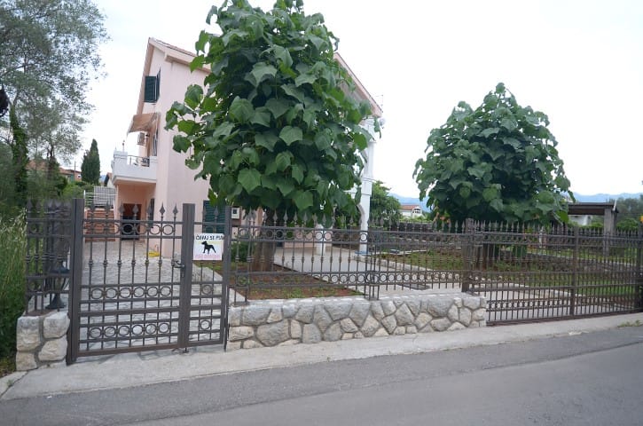 Two-story house in Krašići, Montenegro, with a fenced plot, stone fence, and green trees in the yard.