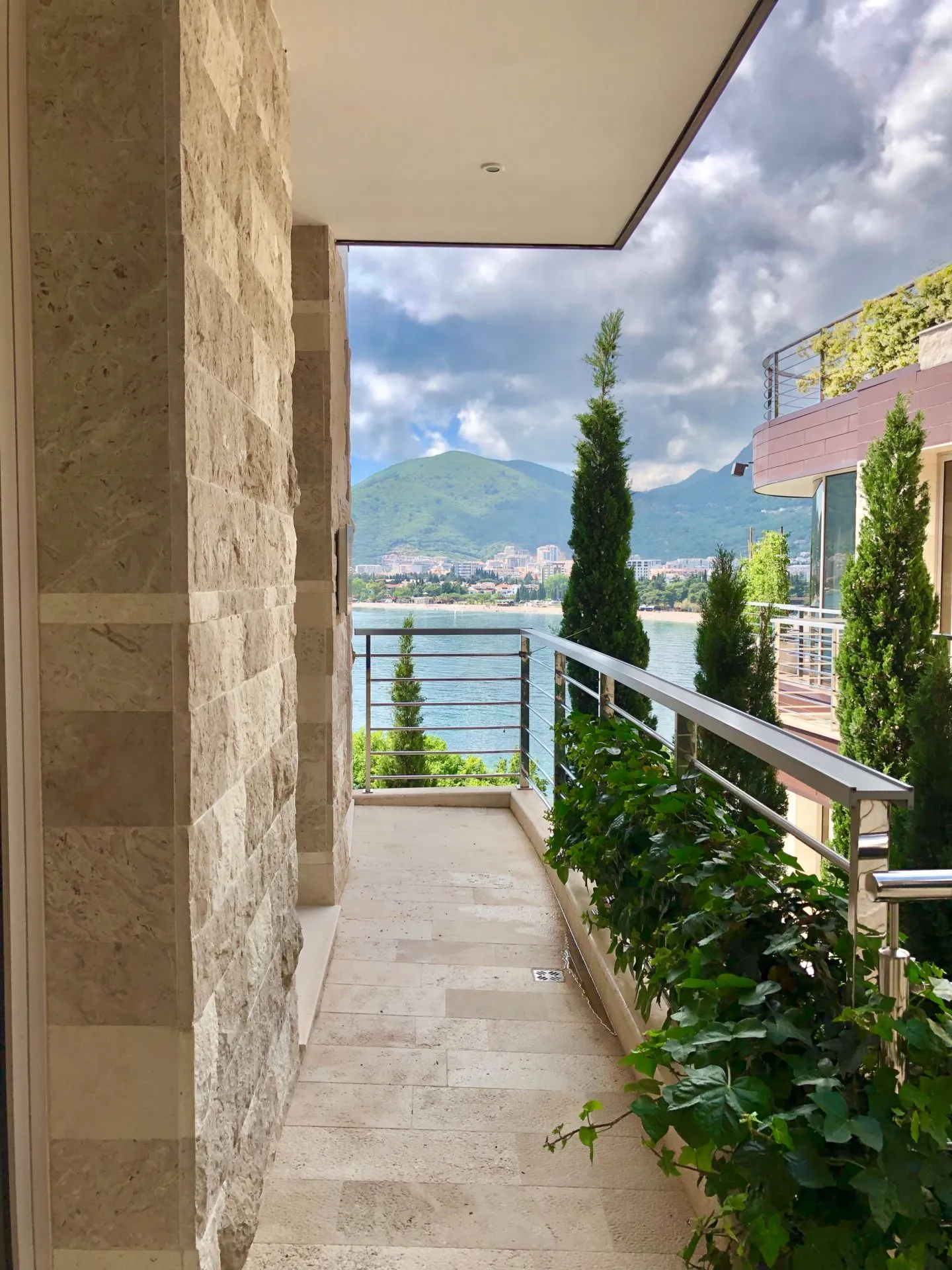 Stone balcony with metal railing overlooking the sea and mountains. Landscaped greenery and tiled flooring visible.