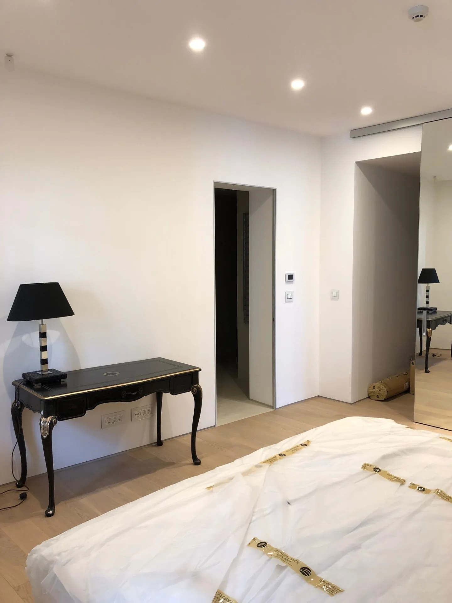 Bright bedroom with white walls, light wood floors, a black console table, and a mattress wrapped in protective plastic.