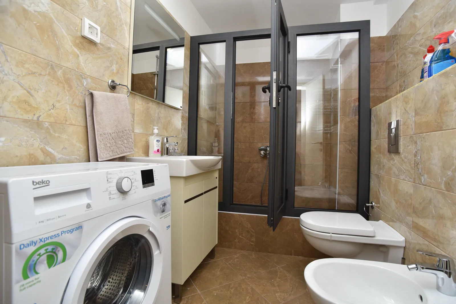 Bathroom with marble tiles, washing machine, sink, toilet, and glass shower enclosure.