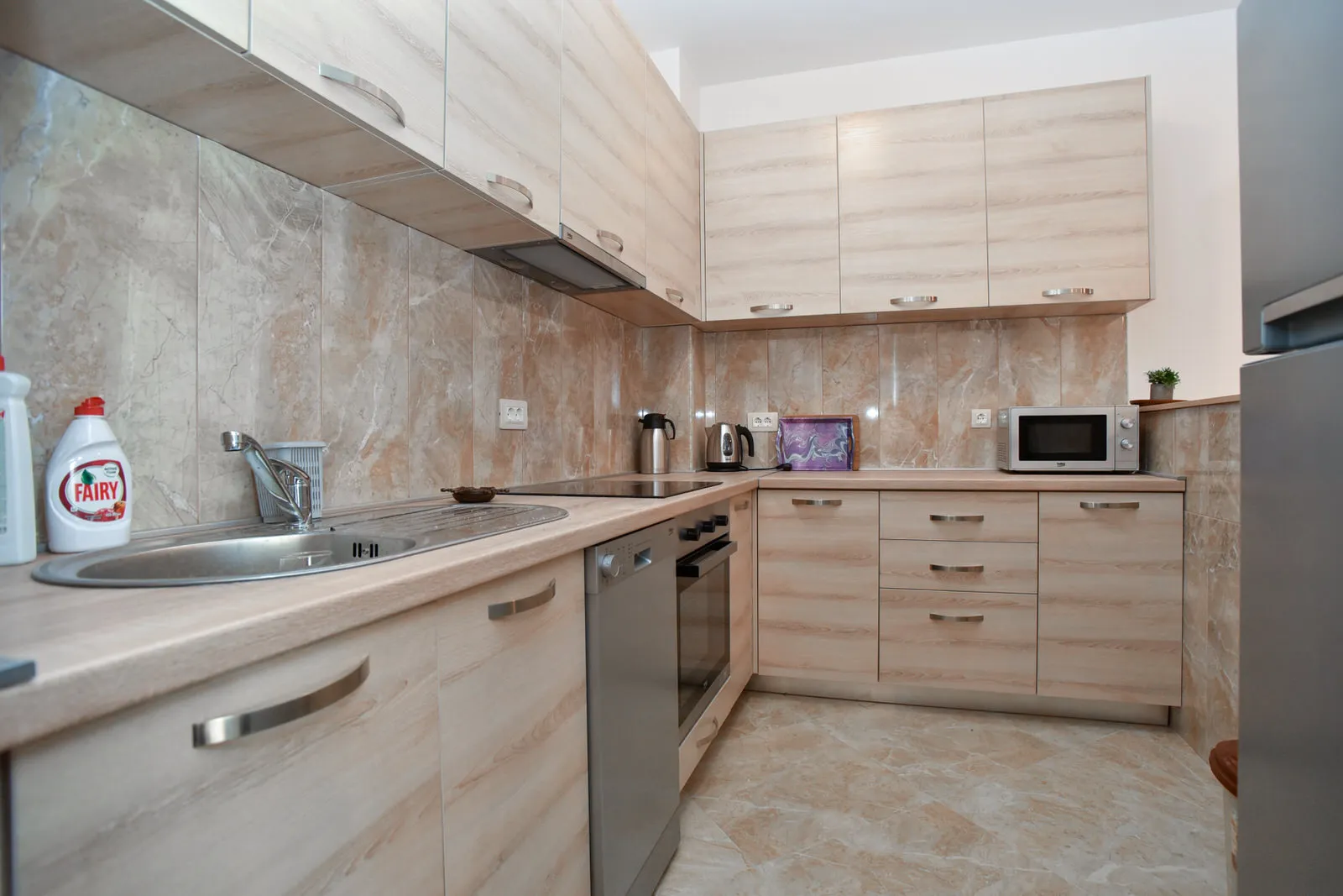 Kitchen with light wood cabinets, sink, dishwasher, microwave, stove, and marble-style backsplash.