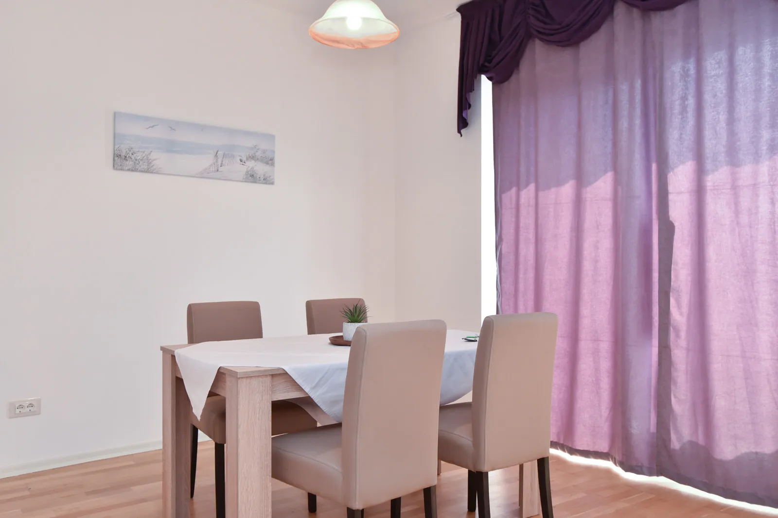 Dining area featuring a light wood table, four beige chairs, and purple curtains against a white wall with a landscape painting.