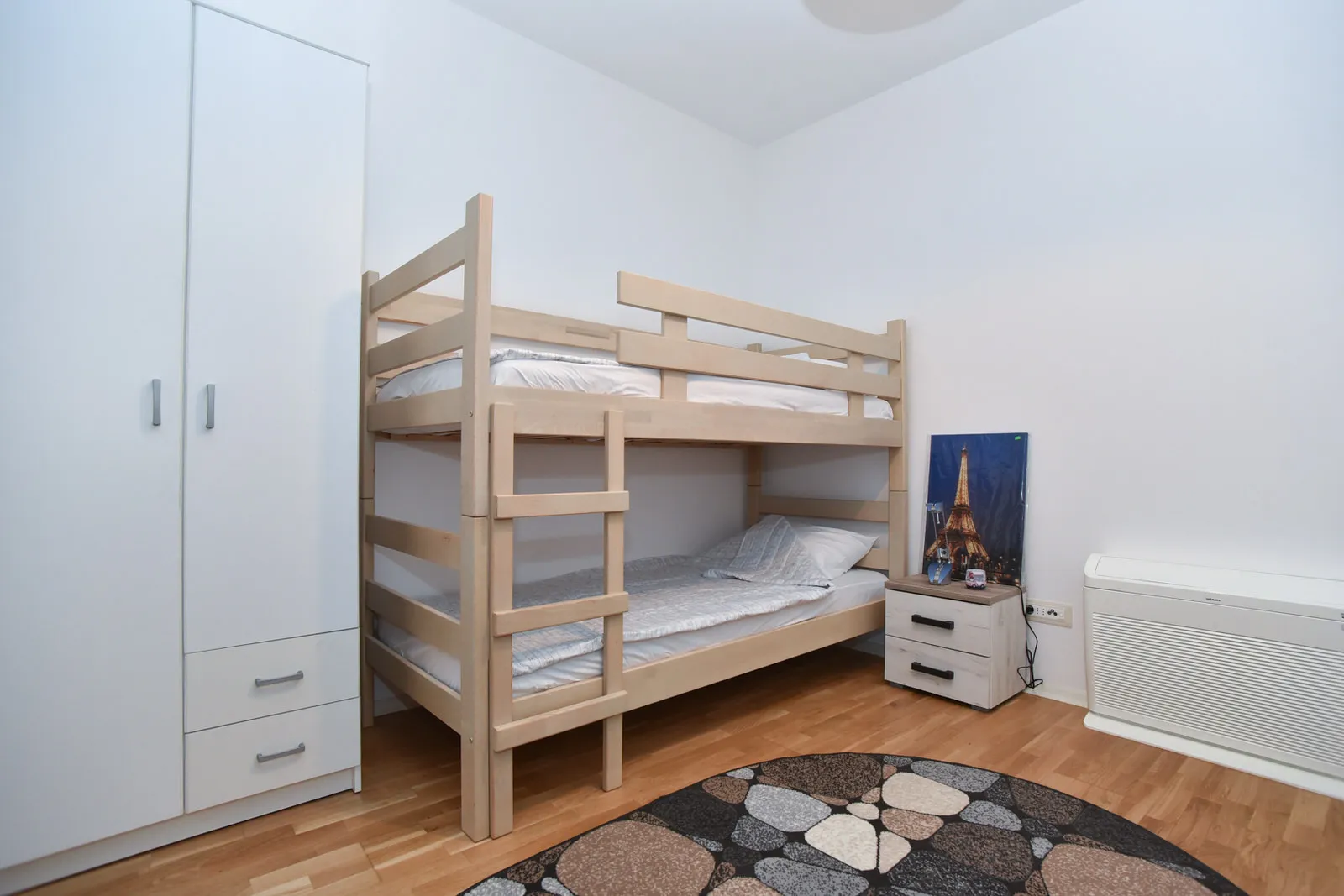 Wooden bunk bed with white bedding, tall white wardrobe, stone-patterned round rug, and nightstand.
