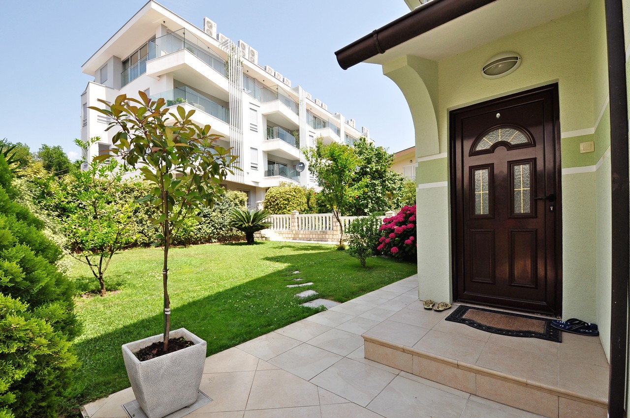 Entrance to a house with a green garden and terrace in Tivat, Montenegro. In the background, a modern residential complex with balconies.