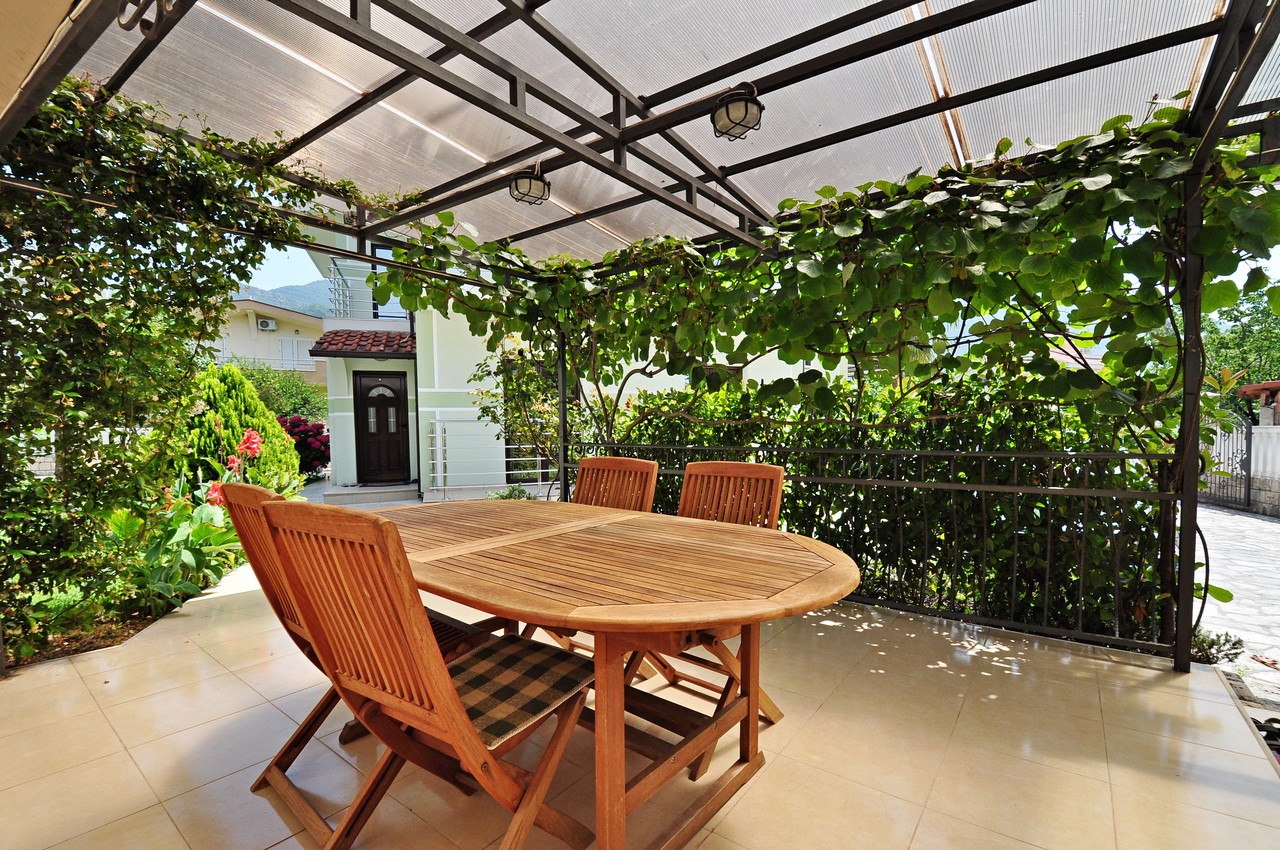 A cozy terrace with wooden furniture and an awning in a private house in Tivat, Montenegro, surrounded by a green garden.