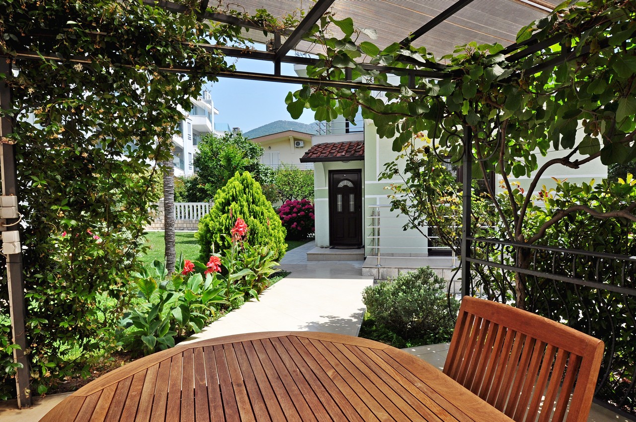 Cozy terrace with a wooden table and a green garden by the house in Tivat, Montenegro, overlooking a well-kept yard.