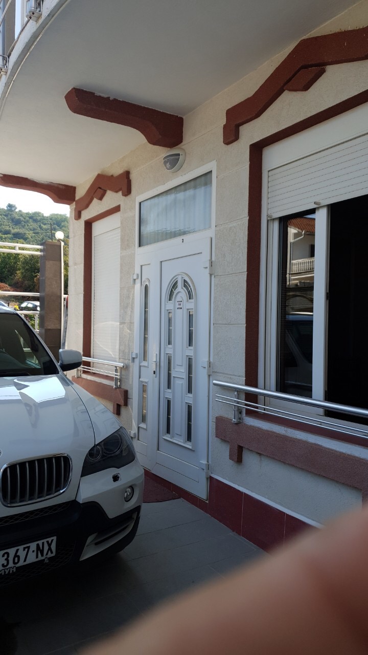 Entrance to a house in Tivat, Montenegro: a white plastic door, windows with roller shutters, and a parking space in front of the facade.