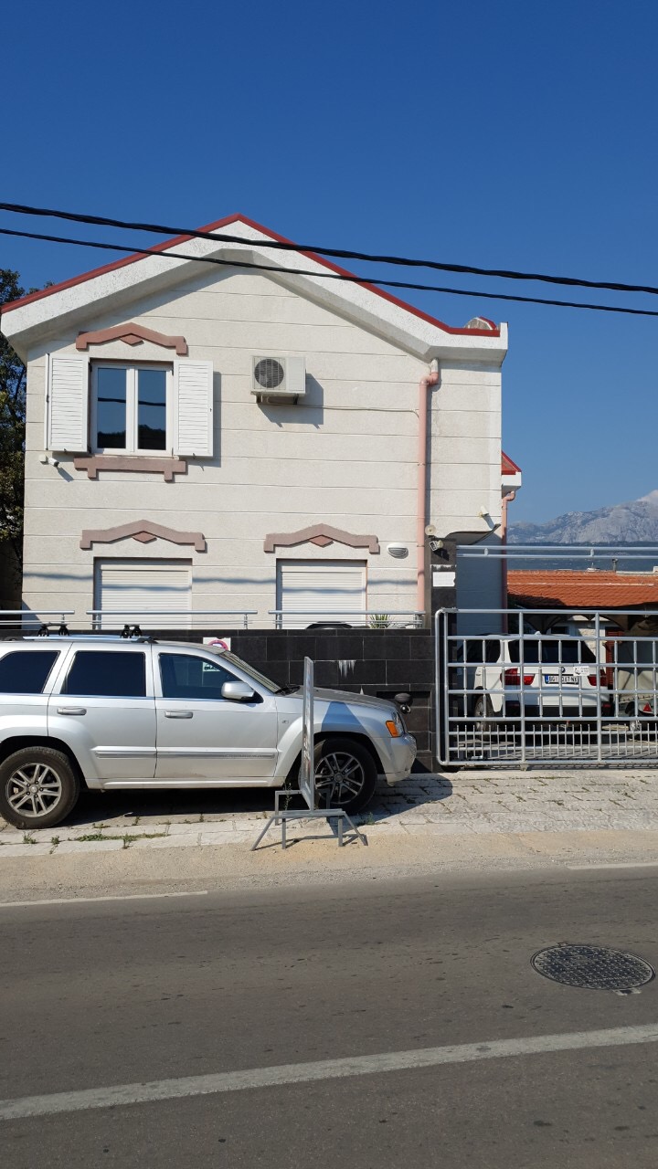 Two-story house in Tivat, Montenegro: facade with light finish, metal gate, and roadside parking.
