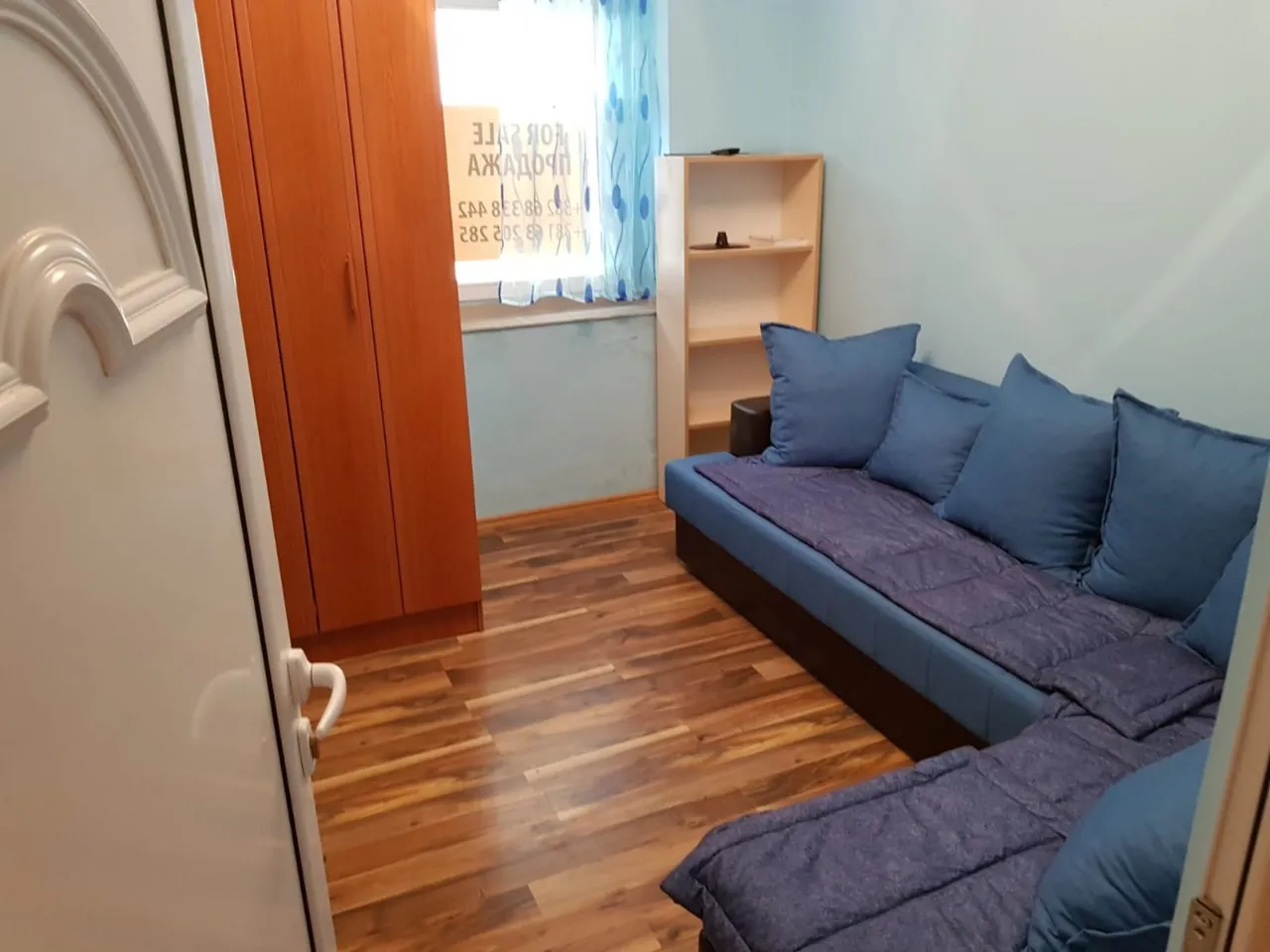 Blue sofa with cushions, wooden wardrobe and open shelving unit near a window with blue curtains.