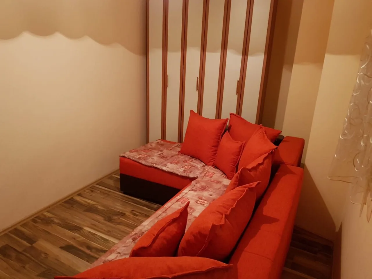 Red L-shaped sofa with cushions and patterned cover in front of a wardrobe and wooden floor.