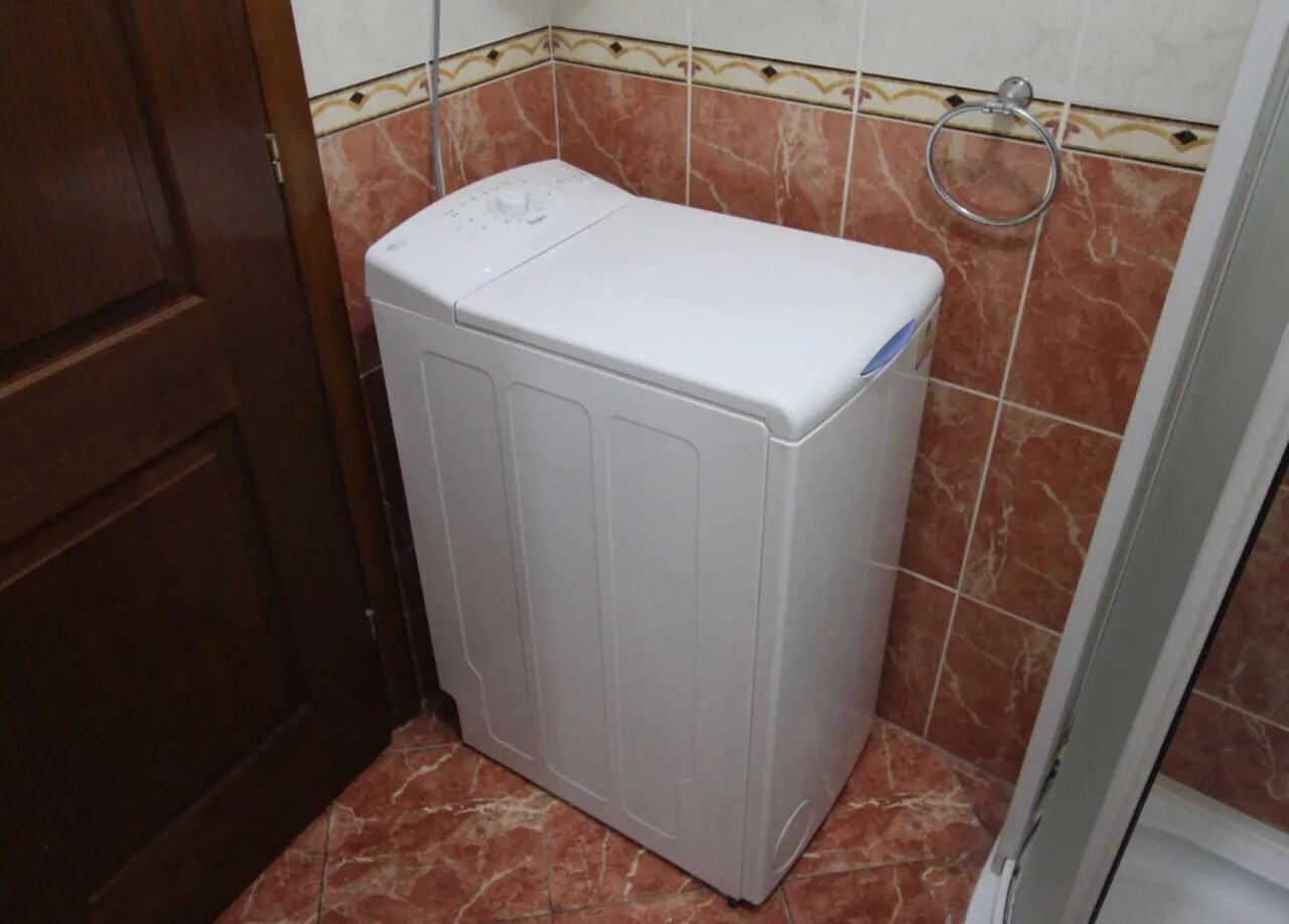 White top-loading washing machine in a tiled bathroom corner next to a towel ring and wooden door.
