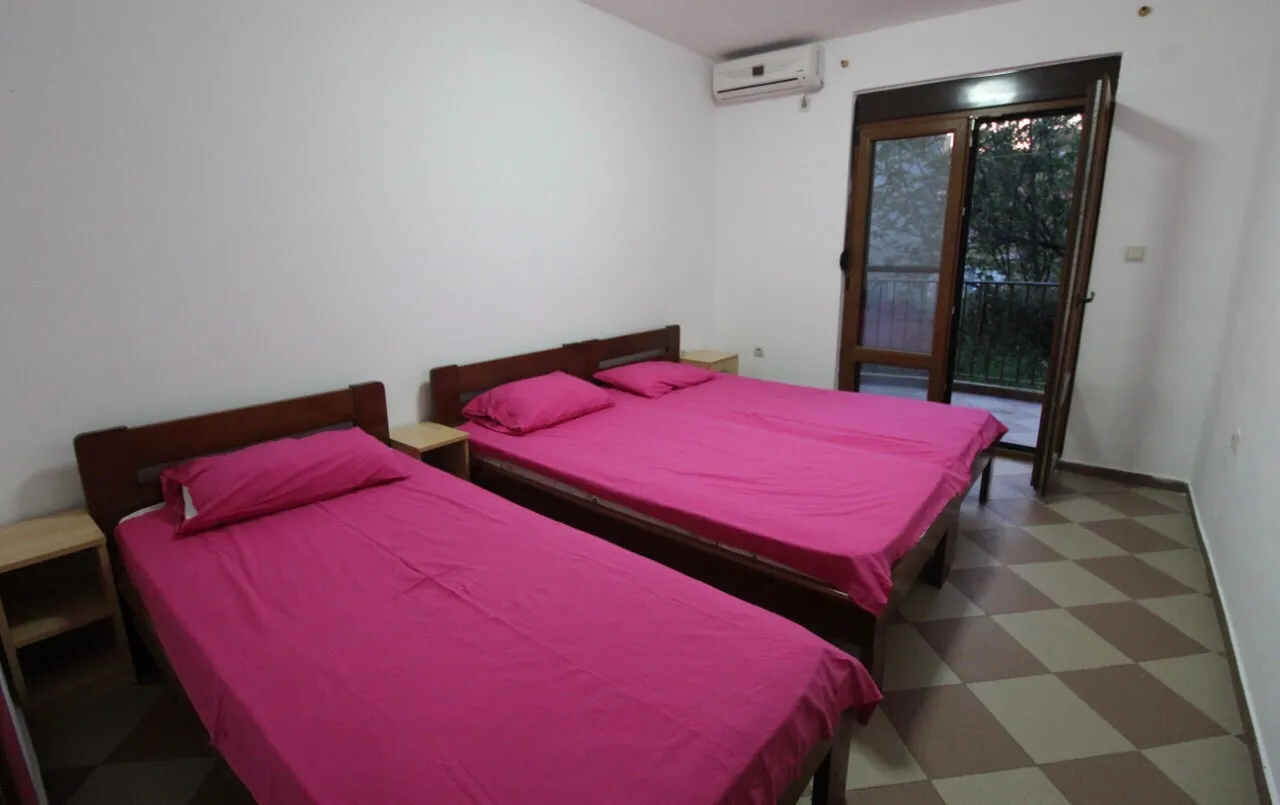 Two single beds with bright pink bedding, wooden headboards, wall-mounted AC unit, and a glass door leading to a balcony.