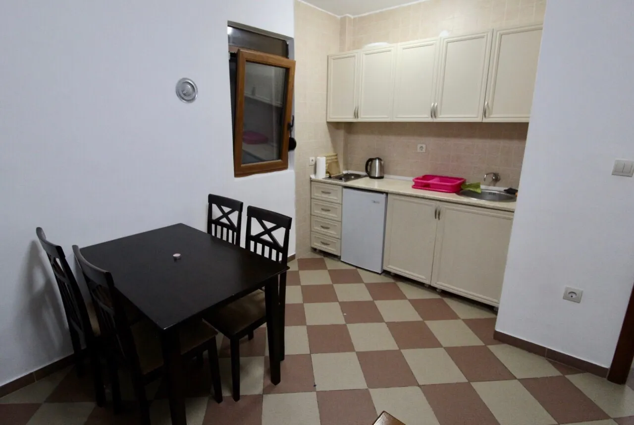 Kitchen with white cabinets, black dining table, four chairs, sink, and small fridge on checkered tile floor.