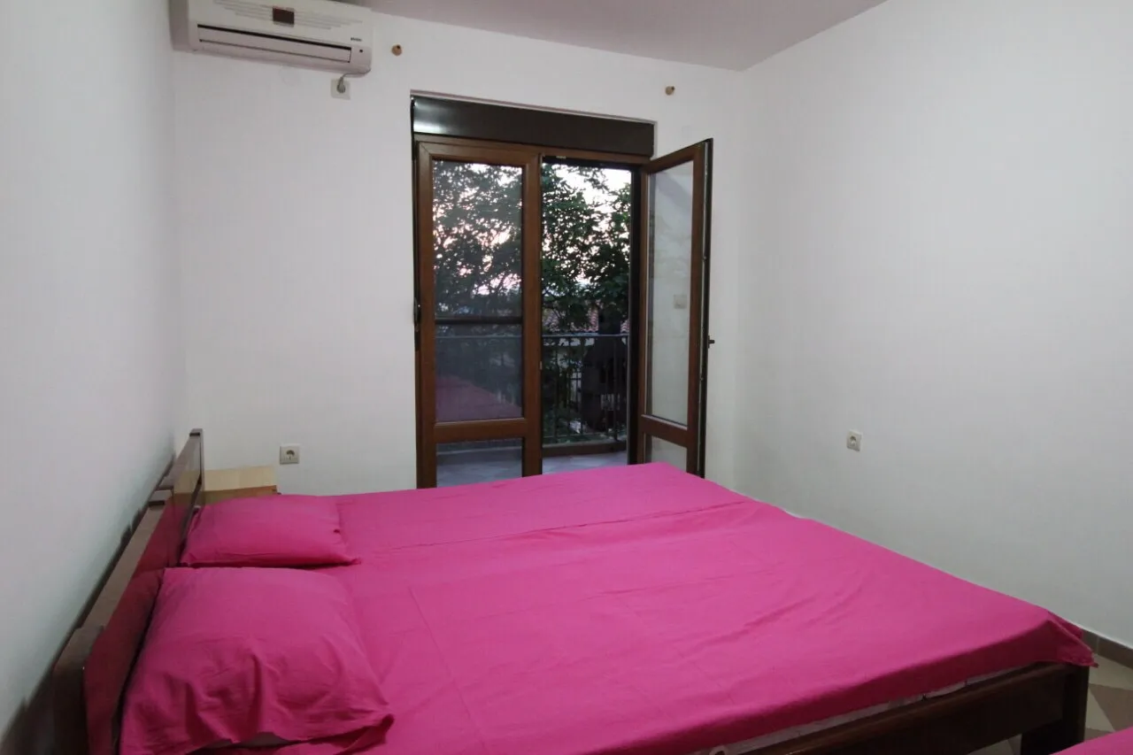 Bedroom with wooden bed and bright pink bedding, wall-mounted AC unit and glass door leading to balcony.