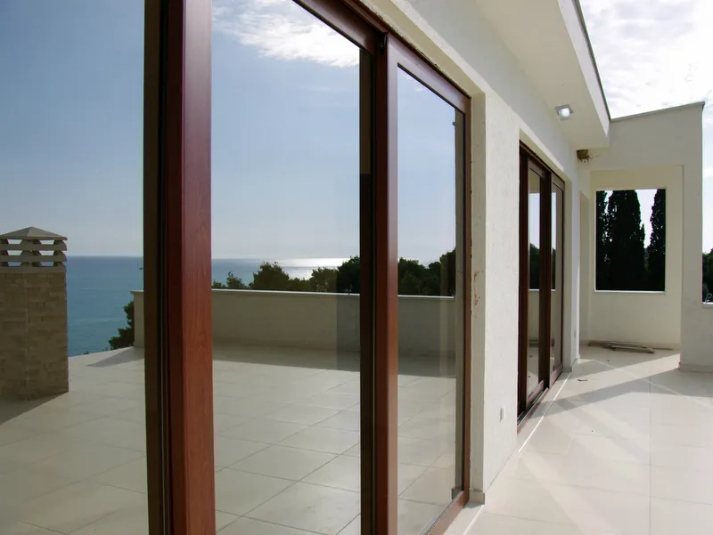 Wide tiled balcony with wooden sliding doors overlooking the sea and trees under a blue sky.