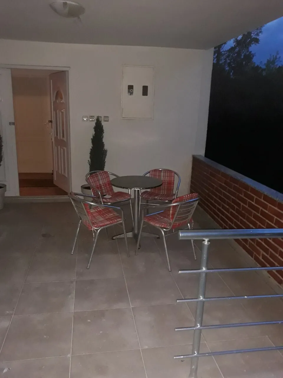 Tiled terrace with metal railing, small round table and four red plaid chairs next to a white wall and open door.