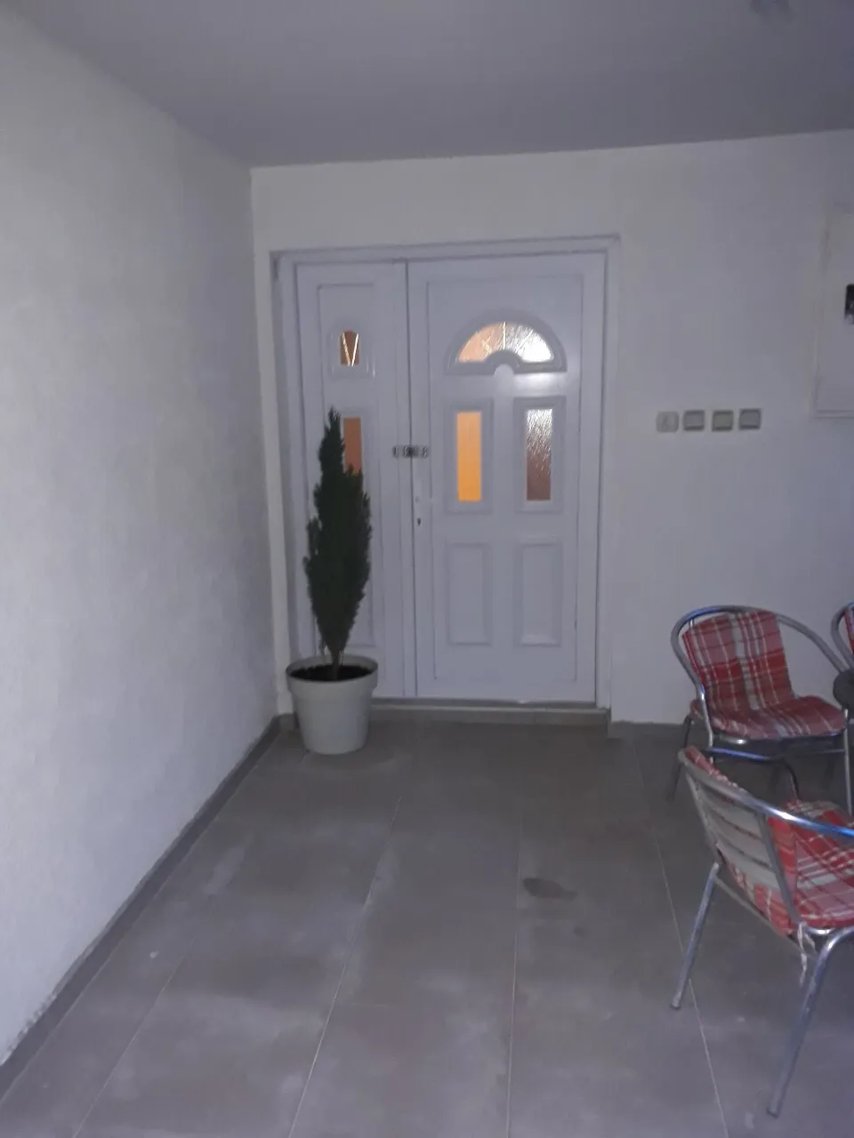 Entryway featuring a white door with glass panels, gray tiled floor, a potted green plant, and two metal chairs with red plaid cushions.