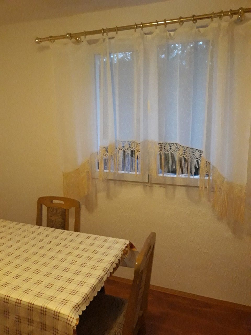 Dining area in the living room of a house in Zhanice, Montenegro: wooden chairs, a table with a checkered tablecloth, and light curtains.
