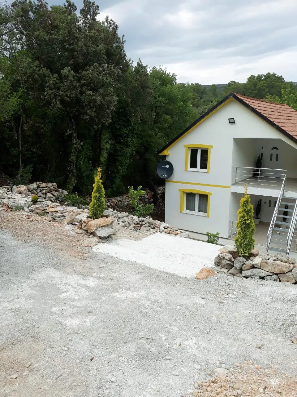 Exterior of a white house with yellow trim, gravel driveway, stone landscaping, and dense trees in the background.