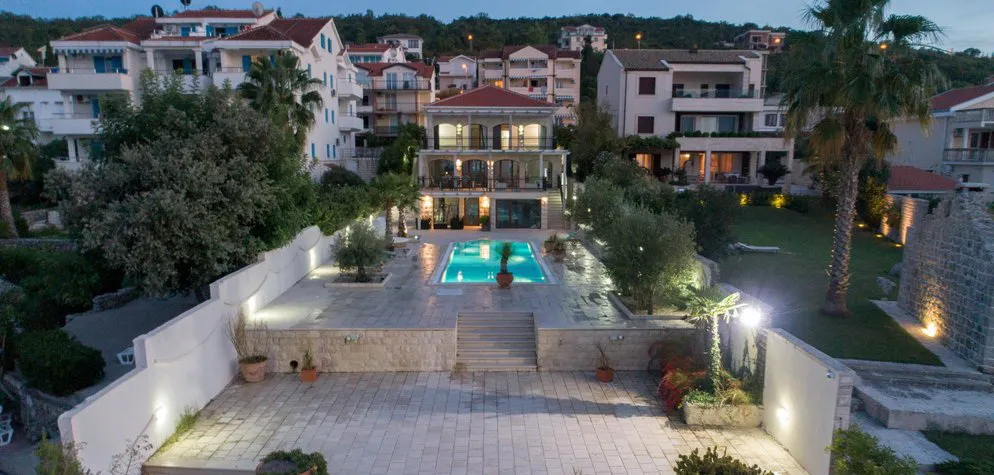 Night view of an illuminated outdoor pool and patio with a villa and surrounding houses in the background.