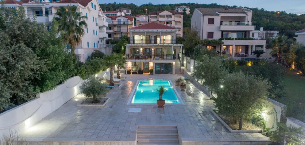Outdoor illuminated swimming pool on a stone patio at dusk, surrounded by trees and residential buildings.