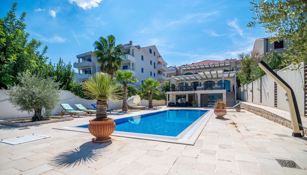 Villa with a pool and lounge area in Krašići, Montenegro: a terrace with sun loungers, palm trees, and a view of a modern house.