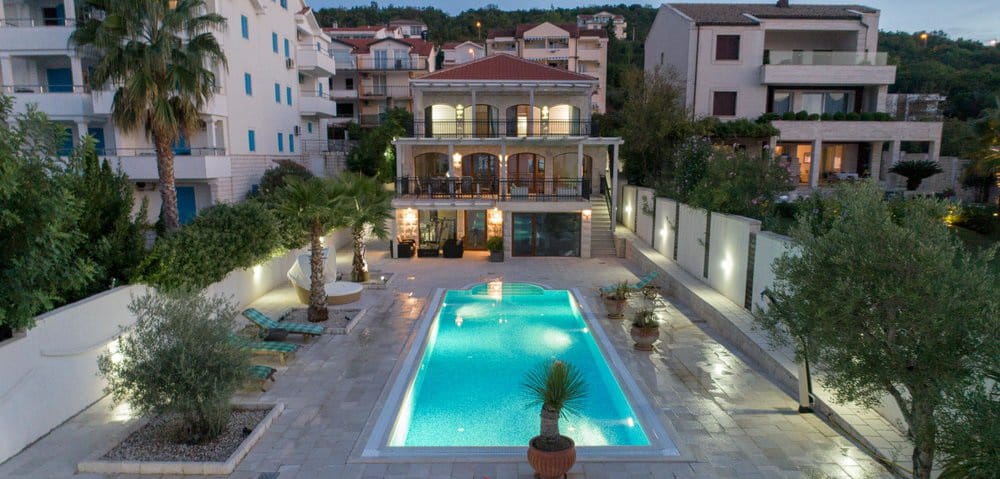Villa in Krašići, Montenegro: view of a three-story house with a pool, terrace, sun loungers, and landscaping in the evening.
