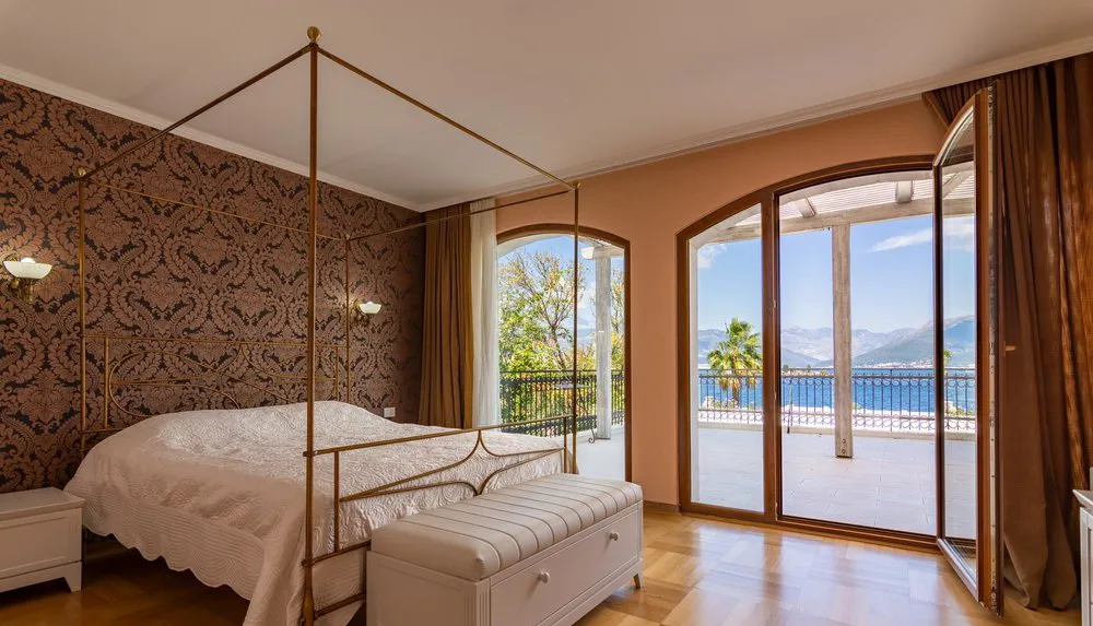 Bedroom featuring a metal four-poster bed, patterned wallpaper, and arched doors opening to a sea-view balcony.
