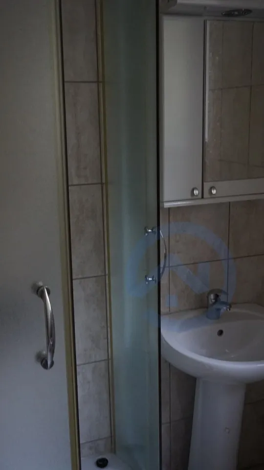 Visible glass shower enclosure with metal handle and white pedestal sink beneath a mirrored cabinet against tiled walls.