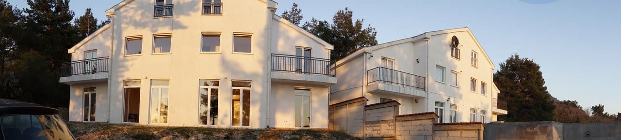White three-story house with balconies in Kavac, Montenegro: modern building facade against a pine forest backdrop.