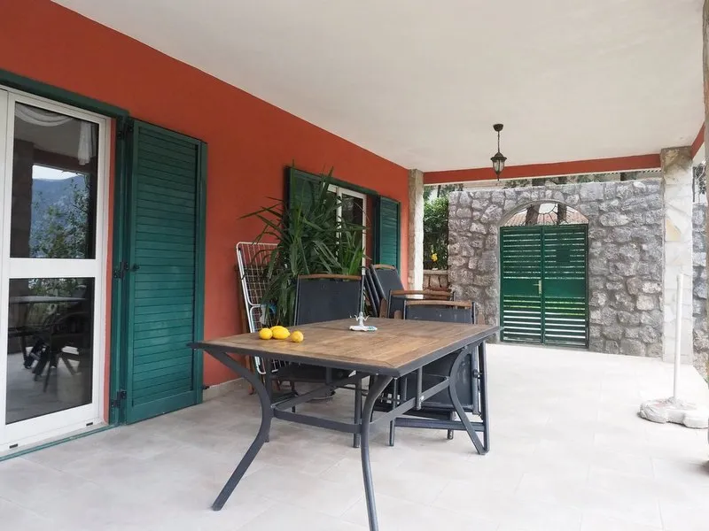 Covered terrace with a wooden table, green shutters, and a stone wall featuring an arched gate.