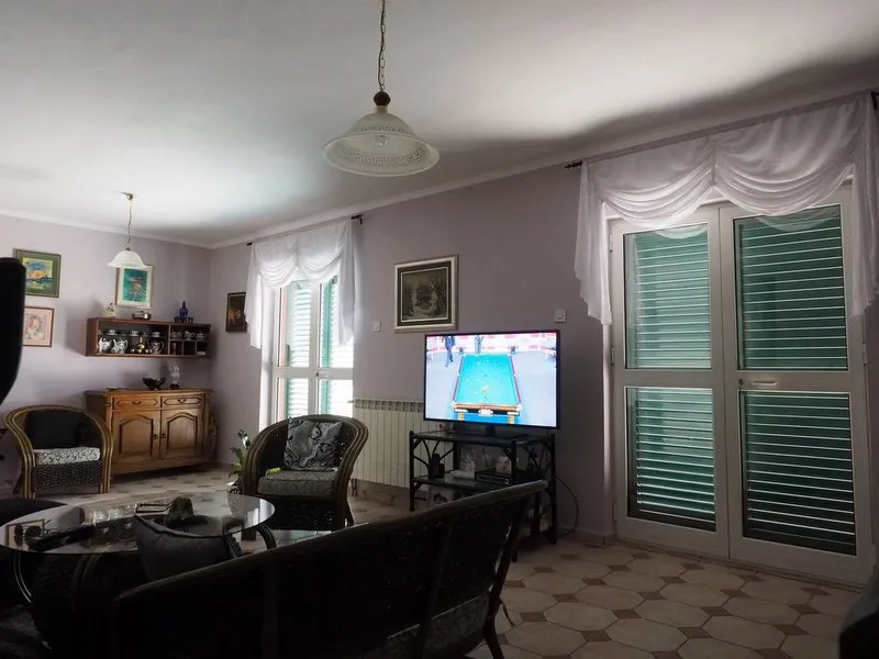 Living room with wicker chairs, TV showing pool game, wooden cabinet, white curtains, and hexagonal tile flooring.