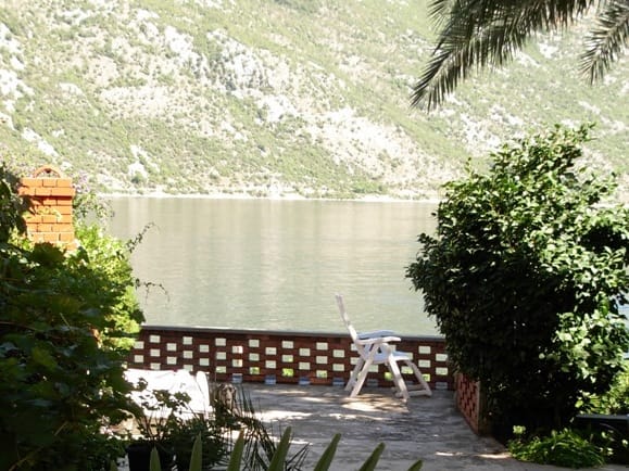 Terrace of a house in Stoliv, Montenegro, with a view of the Bay of Kotor, mountains, and a garden with outdoor furniture.