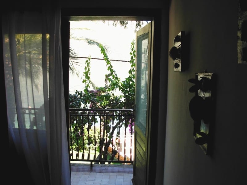View from a balcony of a house in Stoliv, Montenegro: lush greenery, palm trees, and a seascape framed by an open balcony door.