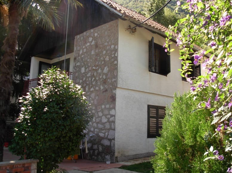 Two-story house in Stoliv, Montenegro: facade with stone finish, tiled roof, and a green garden surrounding it.