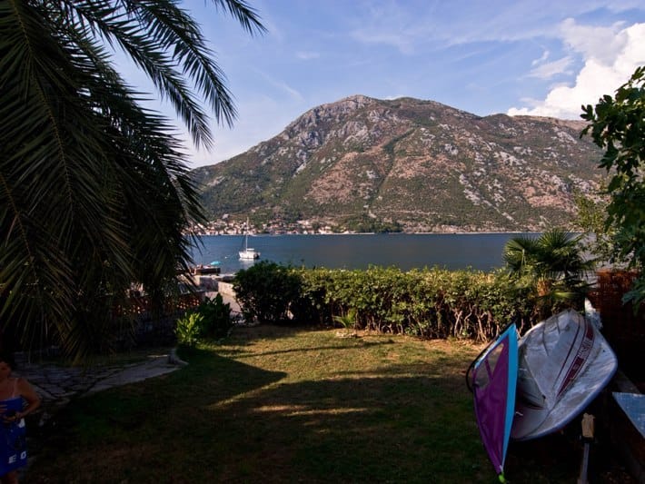 View of the Bay of Kotor and mountains from the garden of a house in Stoliv, Montenegro: palm trees, lawn, and a boat on the seashore.