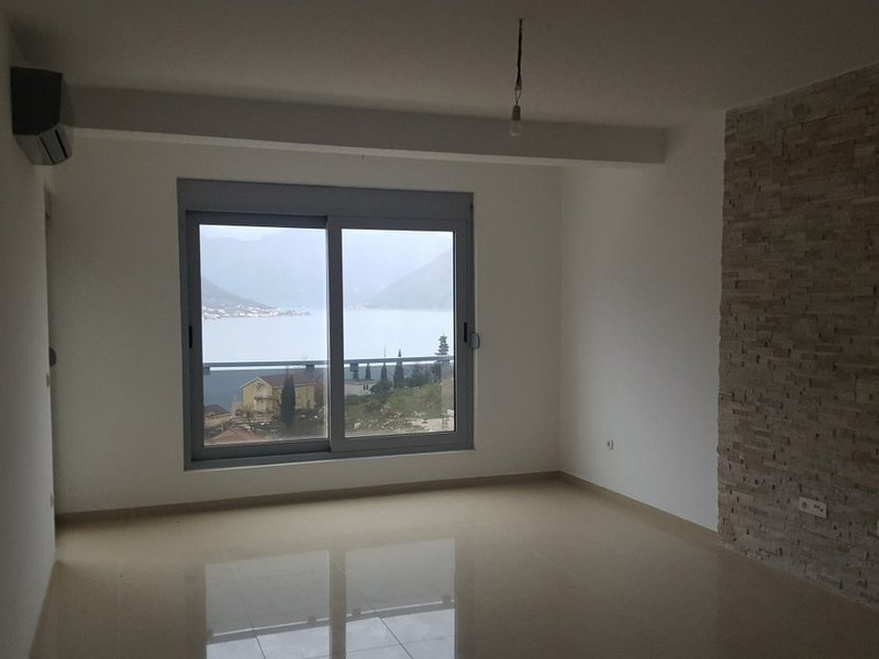 Living room in an apartment in Dobrota, Montenegro: light finishes, stone wall, panoramic window with a view of the Bay of Kotor.