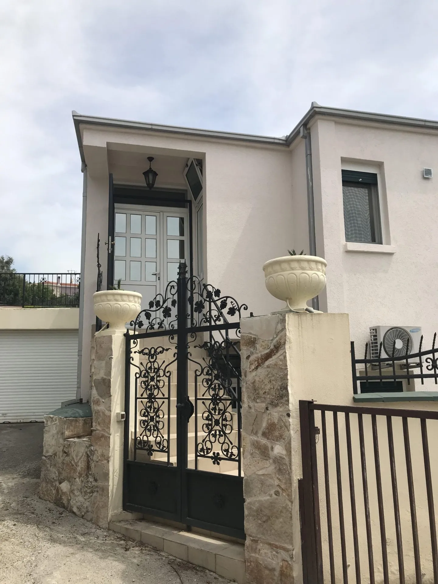 Exterior view of a house facade featuring an ornate black iron gate, stone pillars with white urns, and a cloudy sky.
