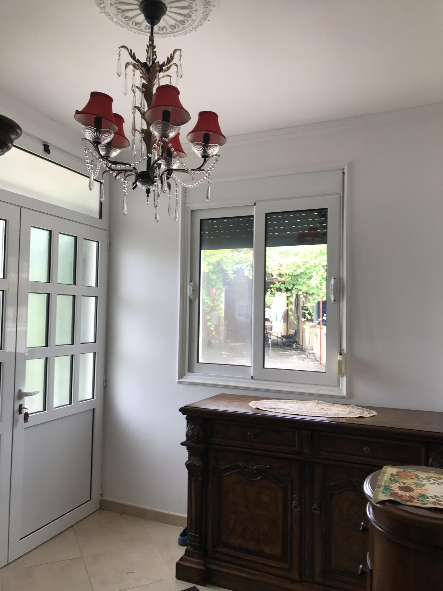 Living room in a house in Bar, Montenegro: an antique dresser, a classic chandelier with red lampshades, and a view of the garden.