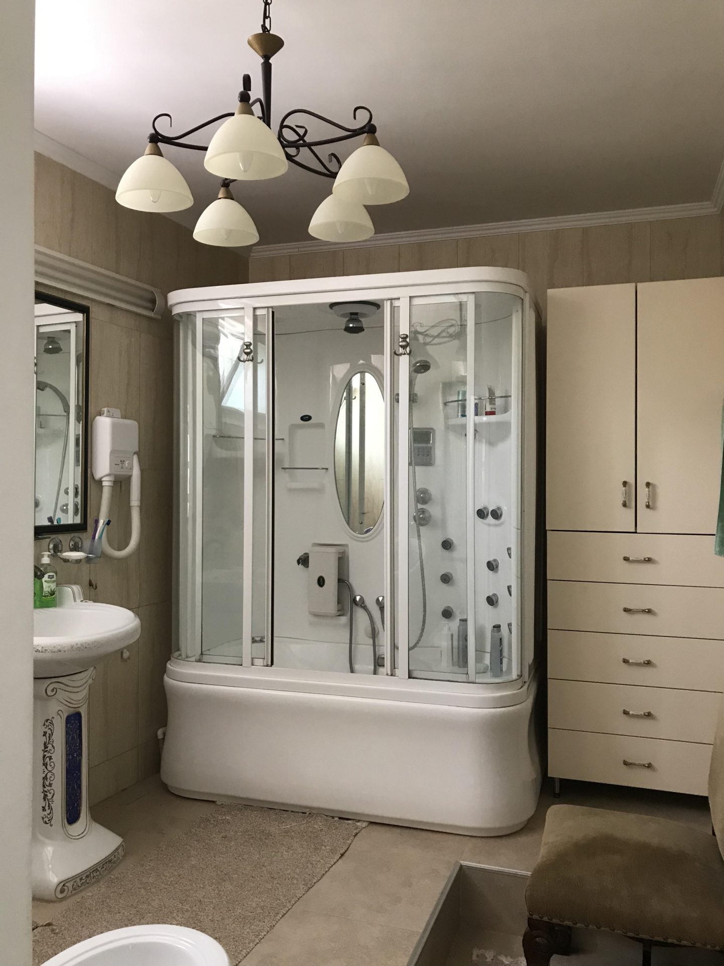 Bathroom in a house in Bar, Montenegro: a hydromassage shower cabin, light-colored furniture, and a classic chandelier.