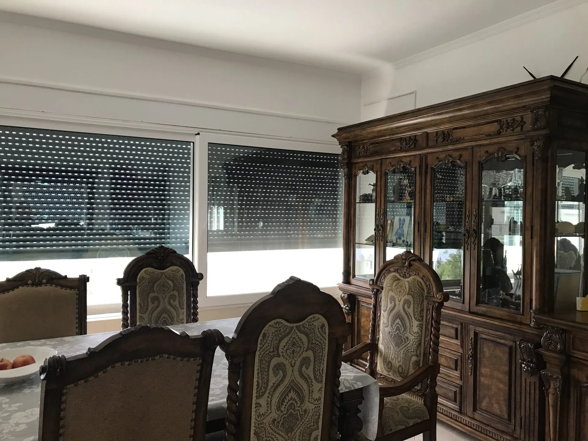 Dining area with a wooden table, ornate carved chairs, and a large glass display cabinet near windows.