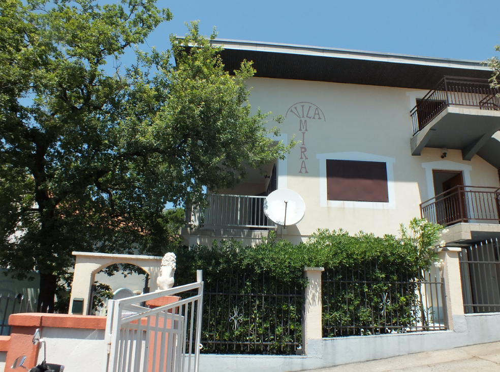 Facade of the two-story Vila Mira villa in Bar, Montenegro, with balconies, a wrought-iron fence, and green trees.
