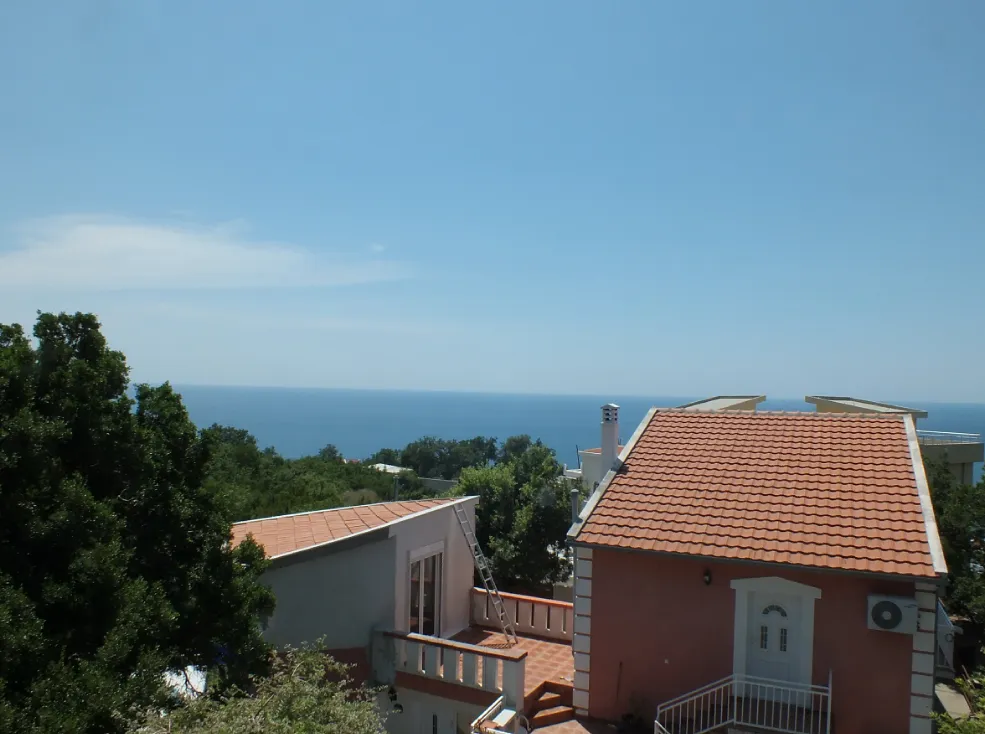 View of houses with terracotta roofs, green trees, and the blue sea under a clear sky.
